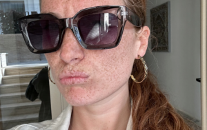 Close-up portrait of a freckled person wearing oversized black sunglasses and a gold hoop earring, with their red hair pulled back in a ponytail.