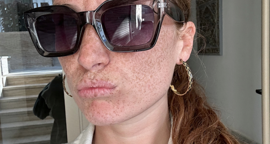 Close-up portrait of a freckled person wearing oversized black sunglasses and a gold hoop earring, with their red hair pulled back in a ponytail.