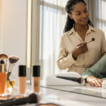 Makeup artist applies powder to a client seated at a marble counter, client has a scab-like scar on the neck near the jawline, mirror behind them.