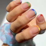 Close-up of a hand with multicolor French manicures on each nail (green, purple, blue, red, orange, yellow) and a blue patterned mask in the background.