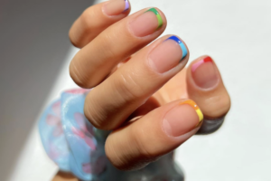 Close-up of a hand with multicolor French manicures on each nail (green, purple, blue, red, orange, yellow) and a blue patterned mask in the background.