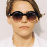 Person wearing oversized round sunglasses with dark gradient lenses, facing the camera against a light wall.