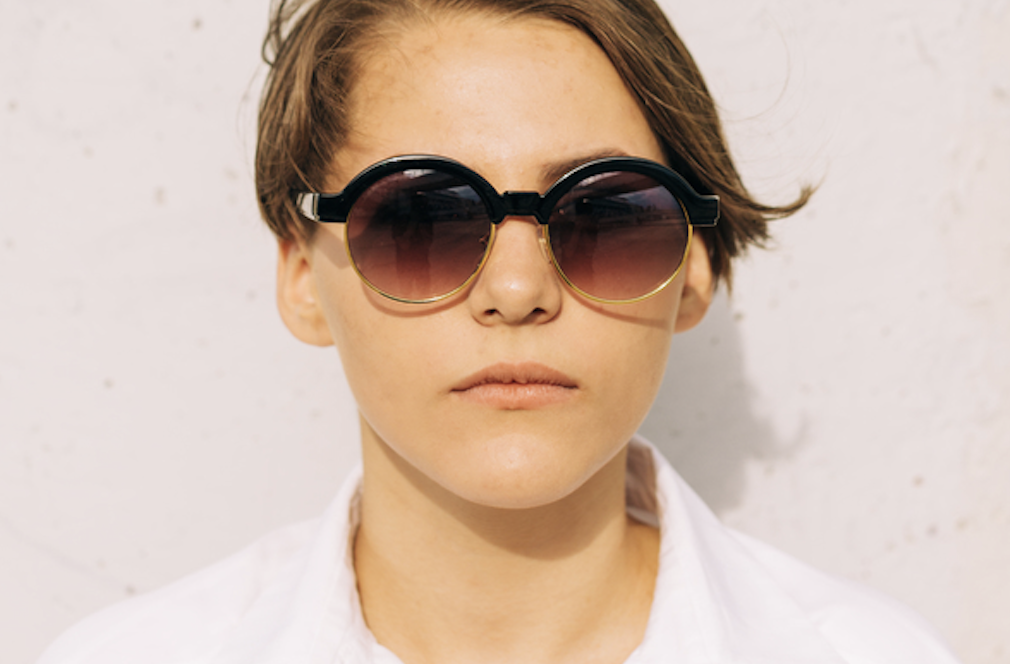 Person wearing oversized round sunglasses with dark gradient lenses, facing the camera against a light wall.
