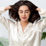 Woman in a cream satin blouse with eyes closed, hands in her hair as she relaxes indoors near green plants.