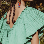 Back view of a person wearing a turquoise-green, ruffled V-back top with braided hair and a colorful beaded earring visible behind the neck in a garden setting.