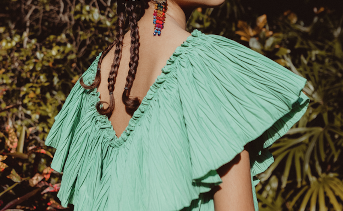 Back view of a person wearing a turquoise-green, ruffled V-back top with braided hair and a colorful beaded earring visible behind the neck in a garden setting.