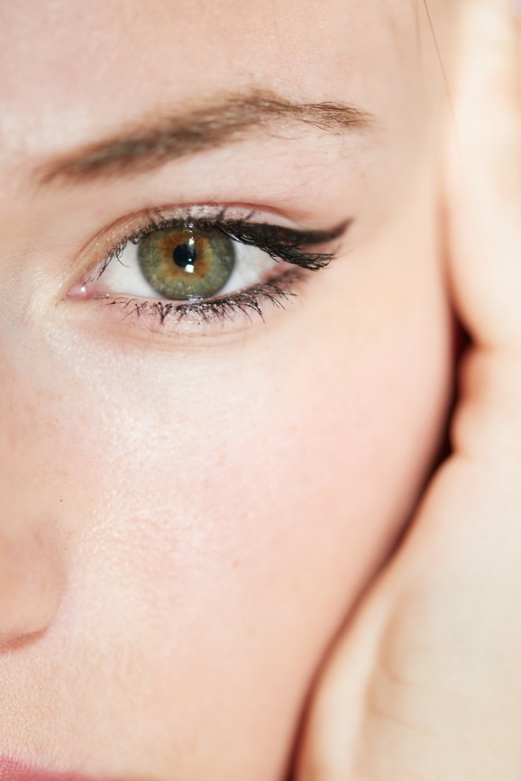 Close-up of a green eye with dark eyeliner, part of the cheek and hand touching the face visible.