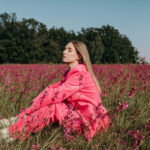 Woman in a bright pink coat sits among pink wildflowers in a sunlit field, eyes closed in peaceful pose.