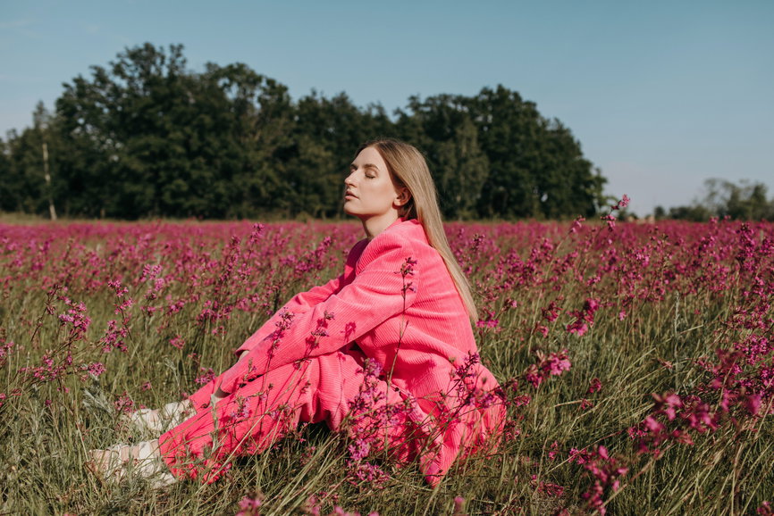 Woman in a bright pink coat sits among pink wildflowers in a sunlit field, eyes closed in peaceful pose.