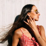 Woman in a red gingham top and sunglasses posing sideways against a light wall, hair blowing in the breeze.