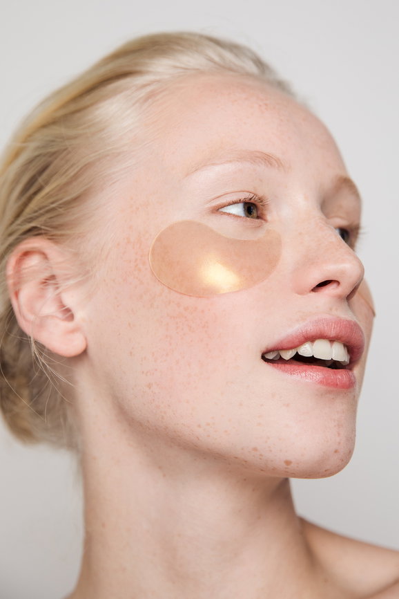 Close-up of a young woman with a beige under-eye hydrogel patch on her left cheek, part of a skincare routine.