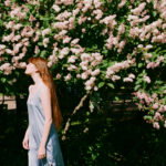 Woman with long red hair in a light blue dress stands in profile beside a bush full of pink blossoms.