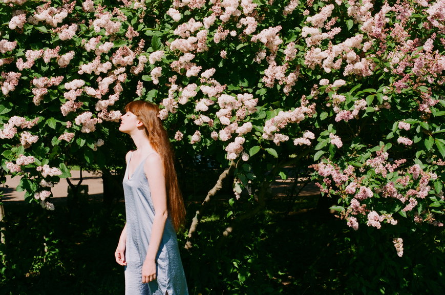 Woman with long red hair in a light blue dress stands in profile beside a bush full of pink blossoms.