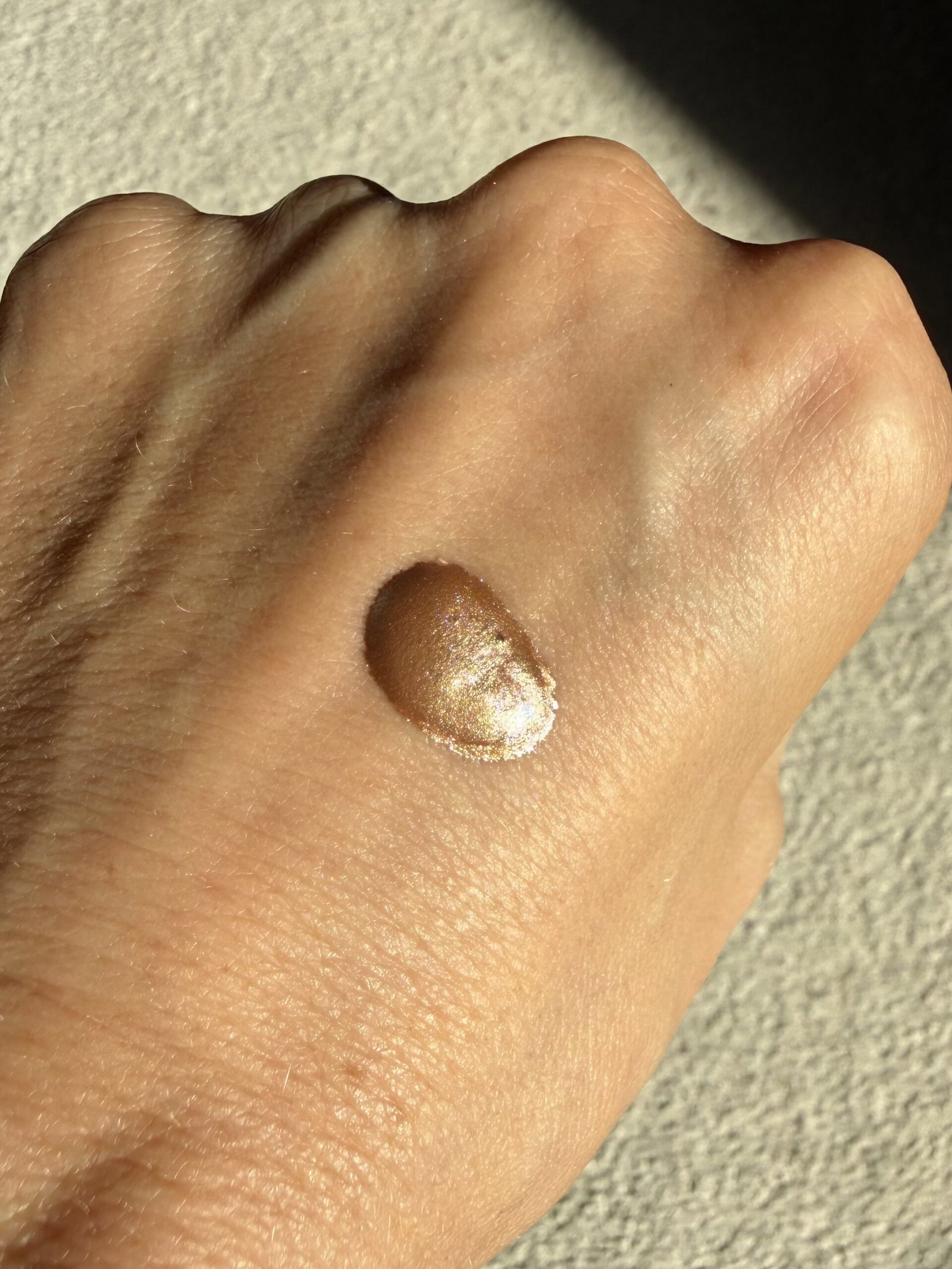 A drop of ICONIC London Illuminator swatched on the back of a hand in direct sunlight, showing its champagne-gold shimmer and metallic finish.