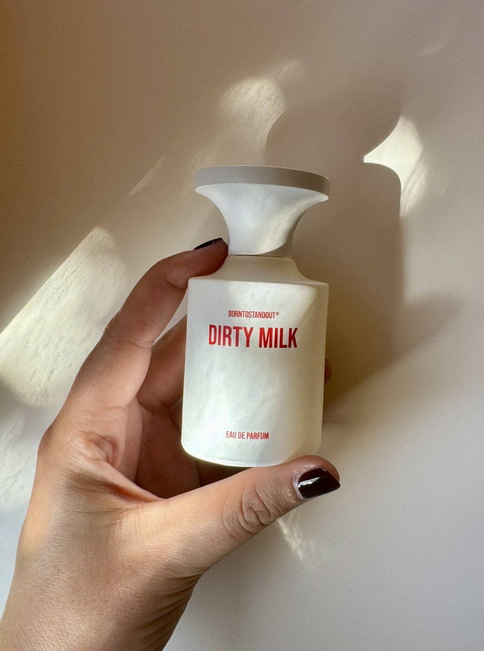BORNTOSTANDOUT Dirty Milk eau de parfum bottle held in hand against a softly lit neutral backdrop.