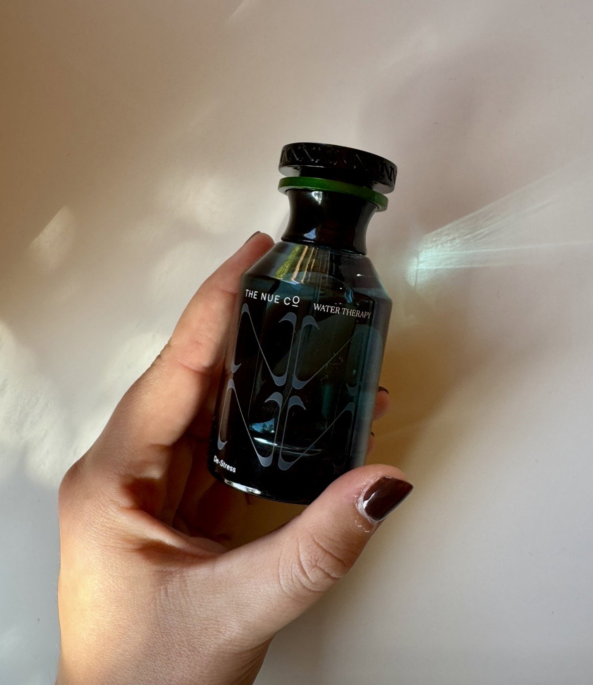 The Nue Co Water Therapy functional fragrance bottle held in hand with sunlight reflecting on a neutral wall.