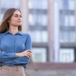 Woman with arms crossed, wearing a blue button-down shirt, standing outdoors in front of a blurred city building backdrop.