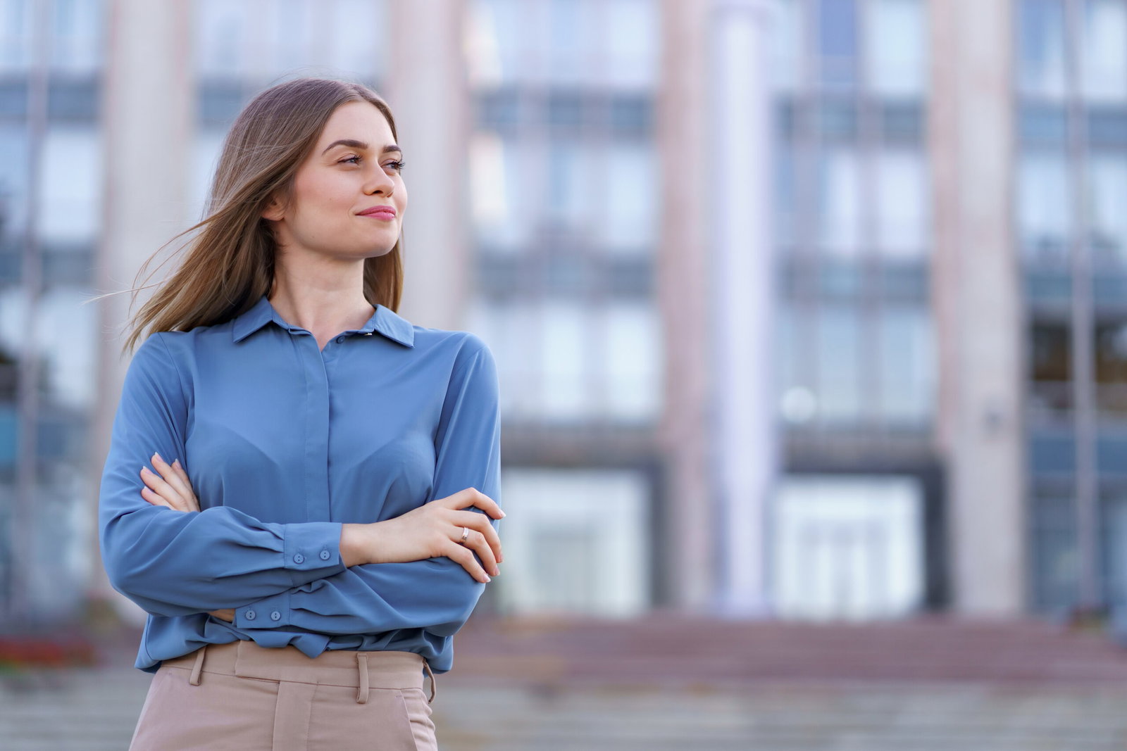 Woman with arms crossed, wearing a blue button-down shirt, standing outdoors in front of a blurred city building backdrop.