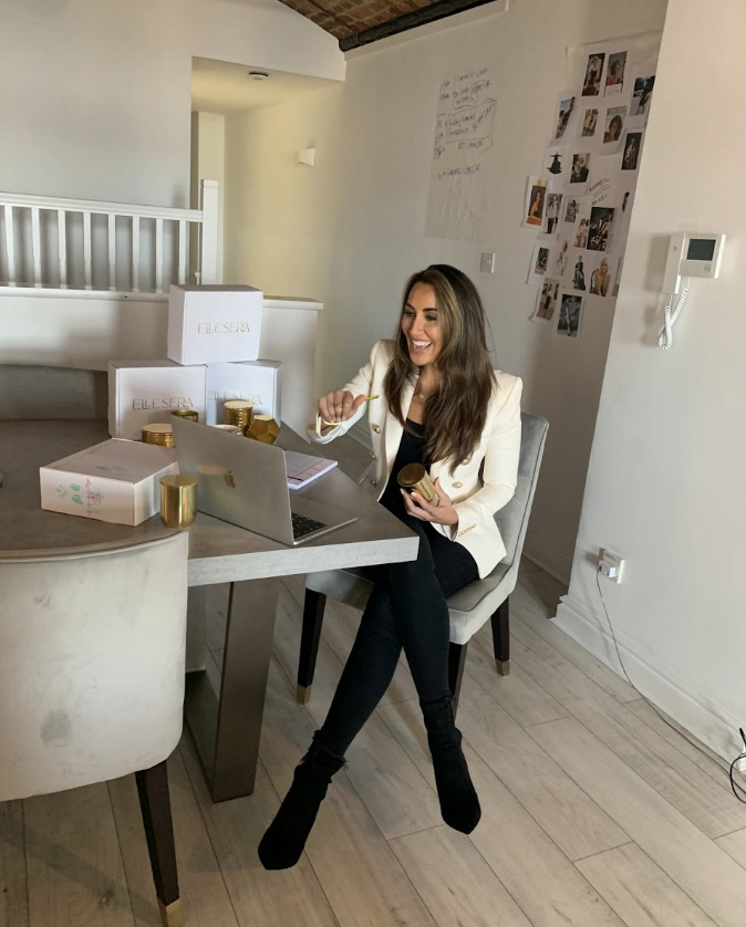 Woman in a white blazer sits at a desk with a laptop, holding a jar and smiling, with ELLE SERA product boxes nearby.