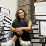 Woman sitting cross?legged among tall stacks of white ELLE SERA boxes in a storage area, smiling at the camera.