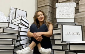 Woman sitting cross?legged among tall stacks of white ELLE SERA boxes in a storage area, smiling at the camera.