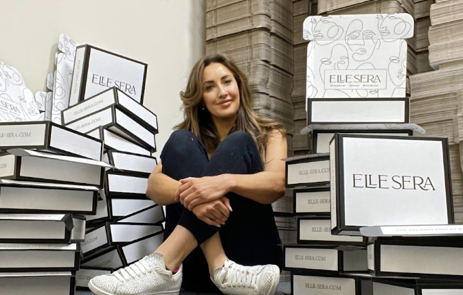 Woman sitting cross?legged among tall stacks of white ELLE SERA boxes in a storage area, smiling at the camera.