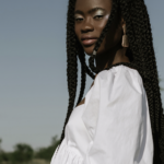 Young woman with long braids wearing a white off-shoulder blouse outdoors on a sunny day.