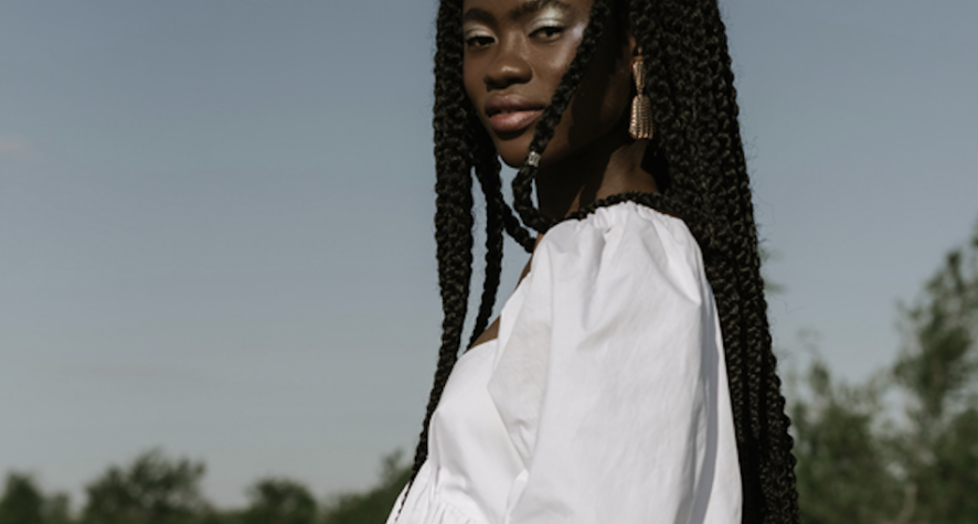 Young woman with long braids wearing a white off-shoulder blouse outdoors on a sunny day.