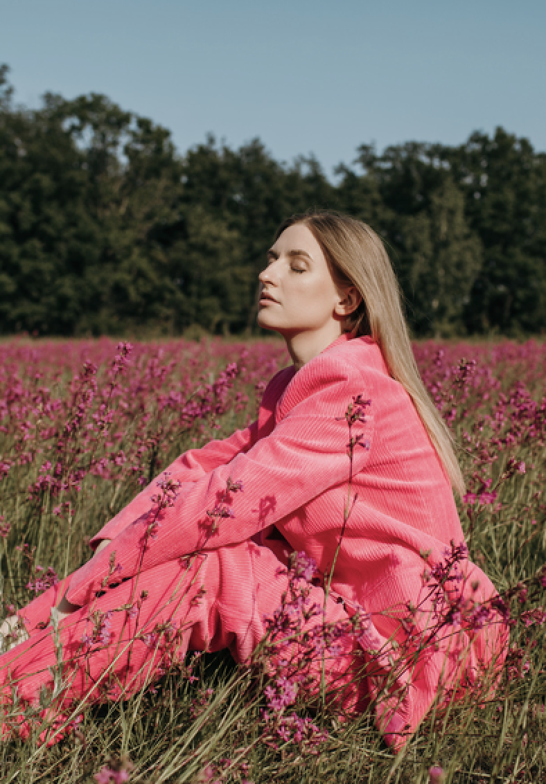 Woman with long blonde hair in a bright pink coat sits in a sunlit field of pink flowers, with trees in the background.