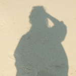 Shadow of a person on pale sand, with one arm raised to touch their head, cast long and dark across the beach.