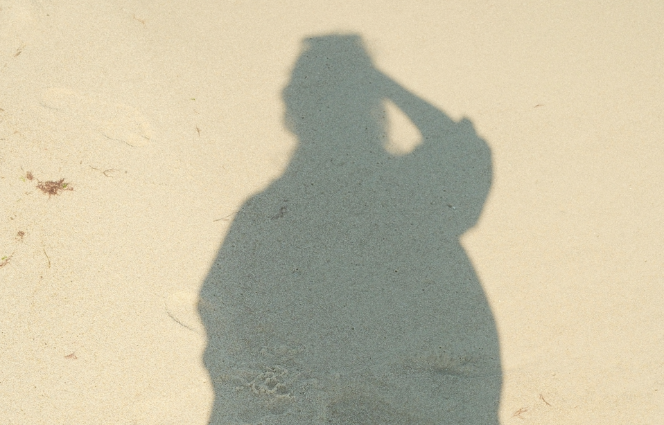 Shadow of a person on pale sand, with one arm raised to touch their head, cast long and dark across the beach.