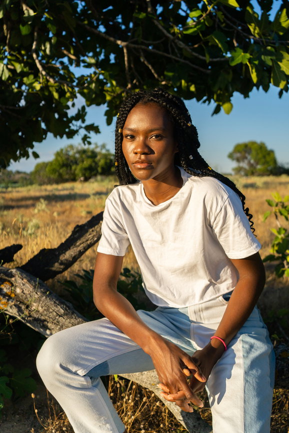 Stock Photo Portrait Of A Young Black Woman On A Tree At Sunset. | Live That Glow Young woman with braided hair sits on a fallen log in a sunny field under a leafy tree