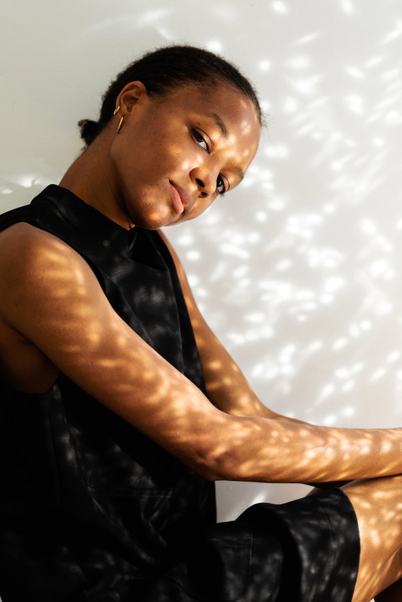 Portrait of a woman in a black sleeveless top sitting against a white wall, with dappled sunlight on her skin.