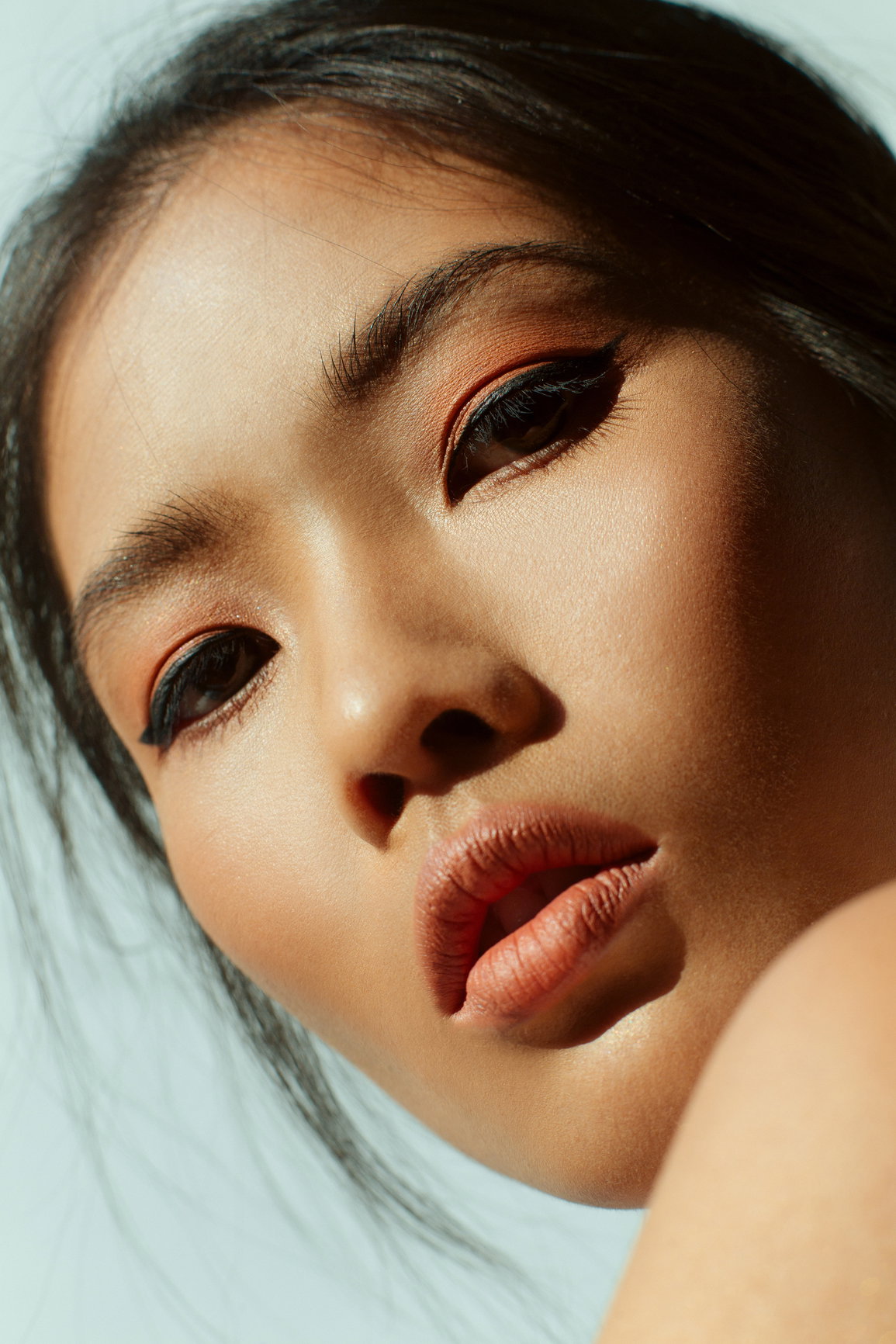 Close-up portrait of a young woman with dramatic winged eyeliner and coral lips, head tilted slightly to the side against a pale blue background.