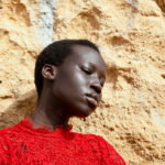 Portrait of a person with short hair wearing a red lace top, leaning against a beige textured wall.