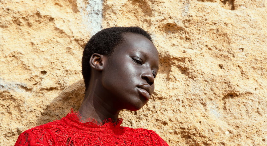 Portrait of a person with short hair wearing a red lace top, leaning against a beige textured wall.
