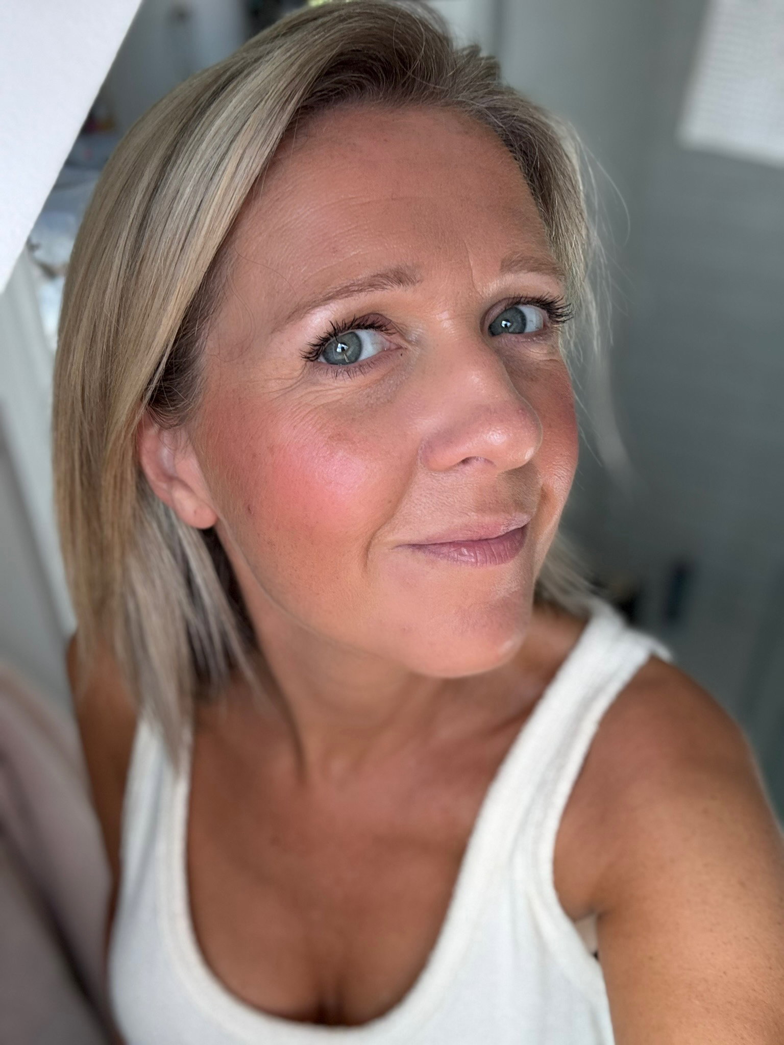 Close-up of a woman with blonde hair and blue eyes wearing a white tank top, looking at the camera.