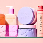 A collection of JOON BYRD skincare and haircare bottles and jars in pastel pinks and purples on a glass shelf against a peach background.