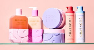 A collection of JOON BYRD skincare and haircare bottles and jars in pastel pinks and purples on a glass shelf against a peach background.