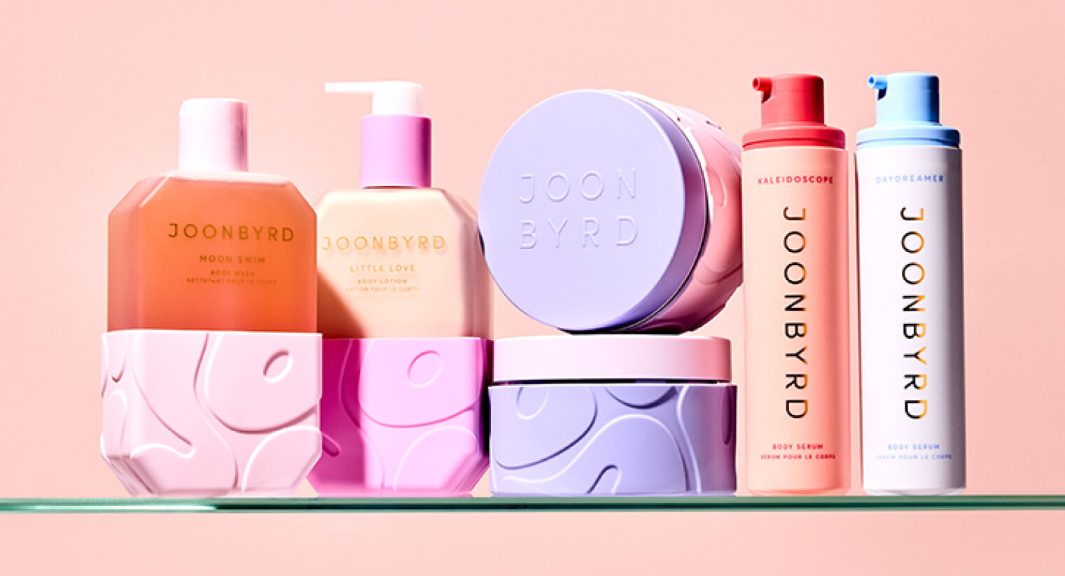 A collection of JOON BYRD skincare and haircare bottles and jars in pastel pinks and purples on a glass shelf against a peach background.
