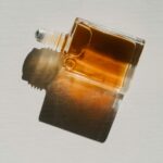 A square glass bottle with amber-brown liquid lies on its side, casting a dark shadow on a light surface.