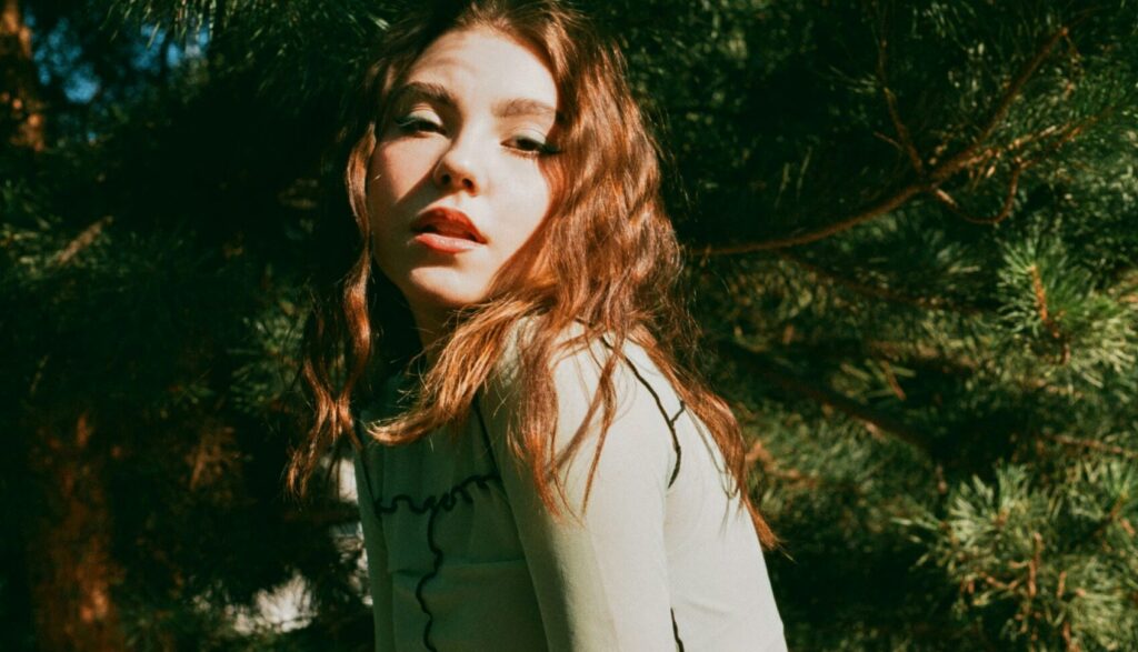 Woman with red hair posing outdoors among evergreen trees in sunlight, looking over her shoulder.
