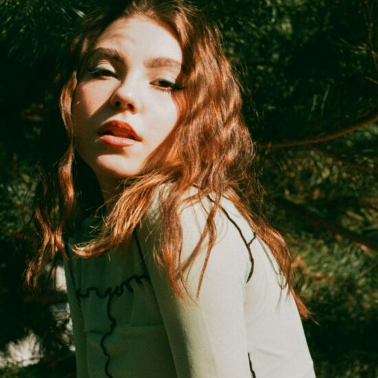 Woman with red hair posing outdoors among evergreen trees in sunlight, looking over her shoulder.