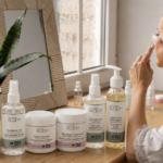 Woman with a towel wrapped around her head applies a facial patch while seated at a wooden vanity with BLOOM skincare bottles nearby.