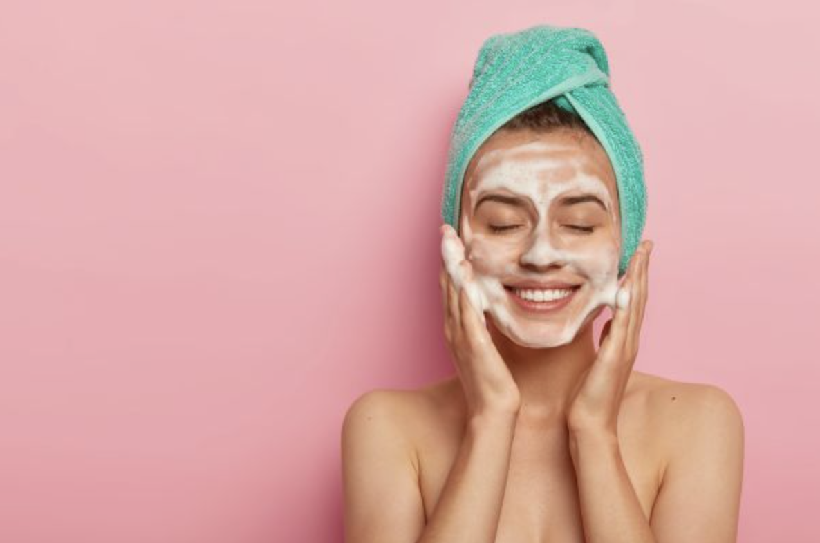 Woman with a white facial mask, a turquoise towel wrapped around her head, smiling with eyes closed against a pink backdrop.