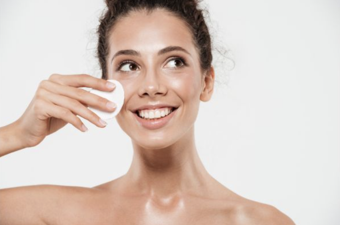 Woman smiling while applying a round cotton pad to her cheek for skincare.