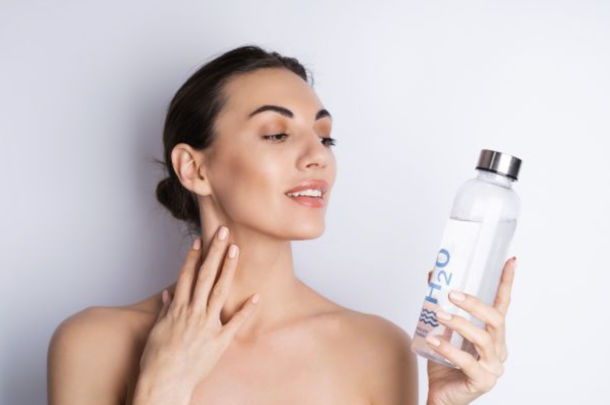 Woman with bare shoulders smiles while holding a clear water bottle and looking at it thoughtfully.