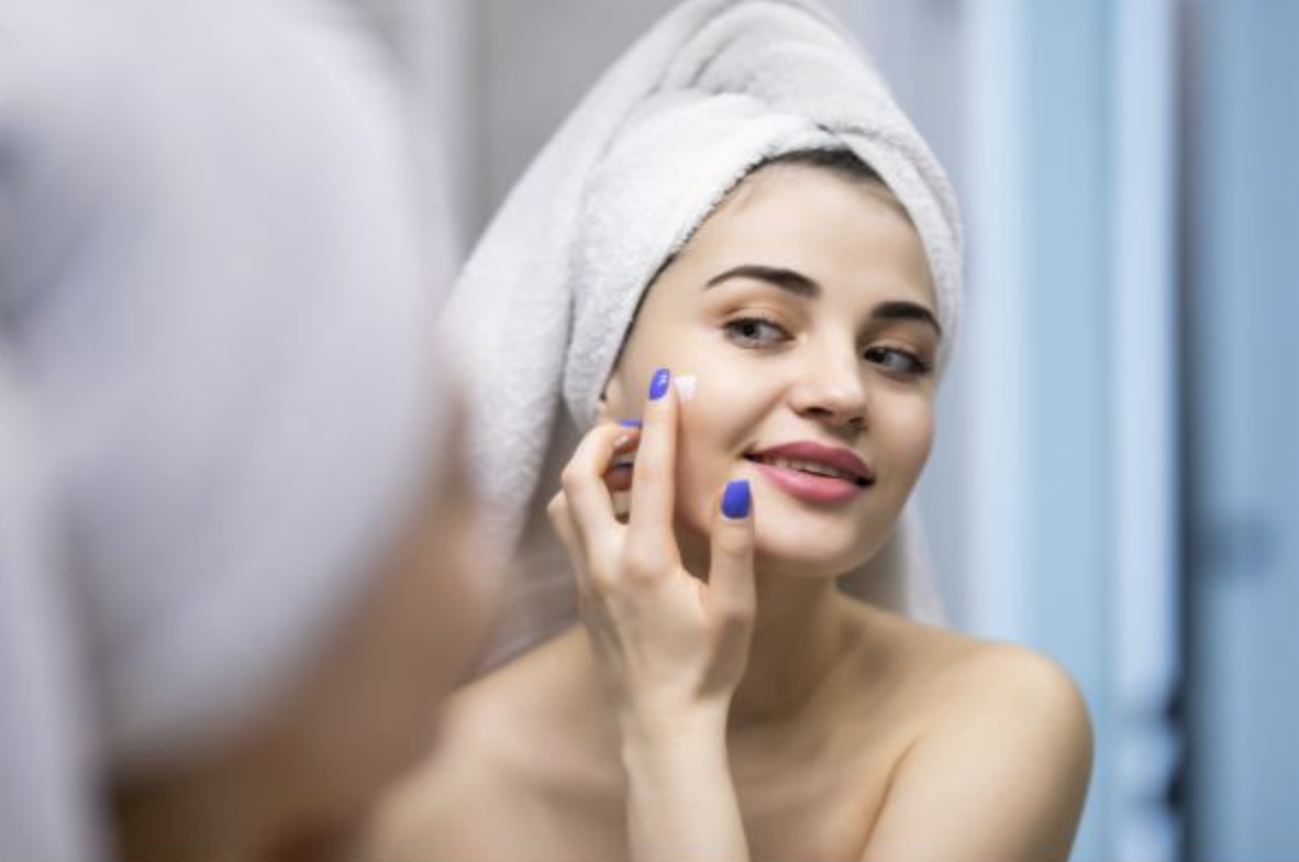 Woman with a towel wrapped around her head applying moisturizer to her cheek in a bathroom mirror, pink lips and blue nails visible.
