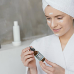 Woman in a white robe with a towel on her head holds a small dropper bottle, applying facial serum in a bathroom setting.