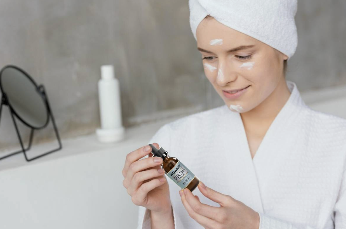 Woman in a white robe with a towel on her head holds a small dropper bottle, applying facial serum in a bathroom setting.